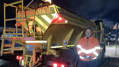 A man dressed in an orange florescent jacket is stood on the right-handside of a yellow gritting vehicle. The man smiles at the camera and has brown hair and a cropped beard and moustache. The gritting vehicle has the word 'spreading' written in red capital letters at the top of its crane. 