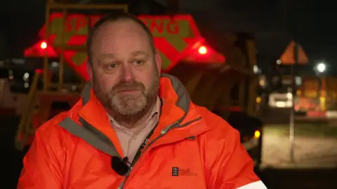 BBC A man dressed in a bright orange jacket with the Hull City Council logo on its right-hand side. He has short brown hair, a cropped beard and moustache and blue eyes. 
