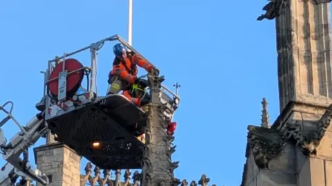 Simon Pattern A person wearing a blue helmet and orange clothing is positioned on an aerial platform above Hull Minster. They are cutting a decorative stone finial on the roof of the building.