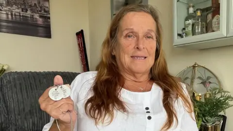 Kevin Shoesmith/BBC Angie Stone, a women in her 60s, with long brown hair, is holding two identification disks, on a chain, stamped with Gregg Stone's Army number, name and religion.