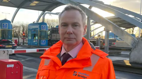 BBC A man with short fair hair looks into the camera as he stands in front of toll booths at the entrance to a bridge. He is wearing a hi-vis orange jacked over a suit, lilac shirt and dark tie. Behind him is a red and white barrier blocking a road. A blue toll booth and red lights can also be seen. A truck is waiting at the toll booth. The sun is low in the sky.