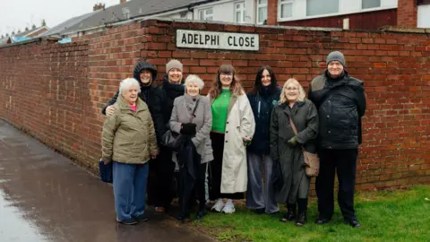 Middle Child/Tom Arran A group of seven women and one man stand in the rain on a residential street. The street sign above their heads reads Adelphi Close.