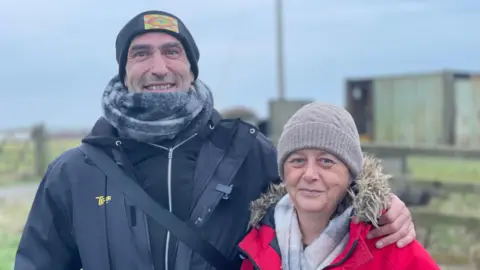 Davey Robinson and Tracy Charlton, Robinson is wearing a black jacket, hat and scarf and has his arm around Charlton, who is wearing a red coat, hat and scarf. Both are looking towards the camera smiling.