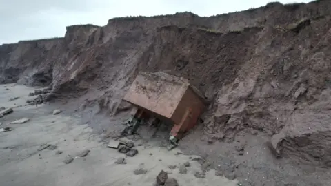 Joe Bilton/BBC Still shot of the bunker at foot of the cliff. It is leaning into the collapsed cliff face and is surrounded by rock and sand.