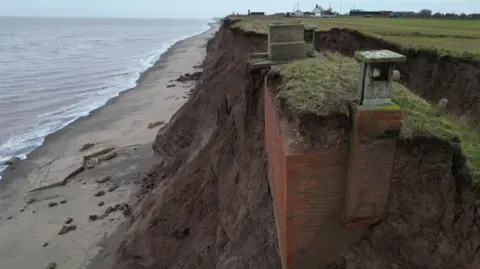 The nuclear bunker - a red brick building - is partially exposed in a muddy clifftop about 20m above a beach. Eroded muddy cliffs are seen in the background. 