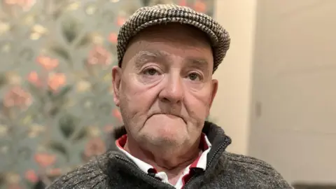 A man in his 60s is sat in his living room. He is wearing a brown check cap, a grey jumper with a zipper at the neck, on top of a red and white striped T-shirt.