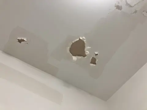 A white ceiling in a bathroom with peeling paint in four areas. The colour of the paintwork is patchy.
