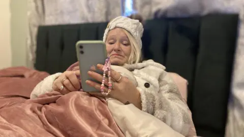 BBC A woman in her 50s is sat in a bed with a pink velvet bedspread and looks at her mobile phone, whch has a grey case. She's wearing a cream-coloured knitted bobble hat and a cream and camel coloured coat. 