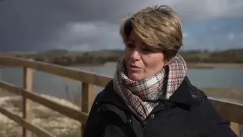 A woman with short brown hair wears a black winter coat and plaid scarf as she stands on a path in front of a wooden fence and a large lake at a nature reserve. The sky is dominated by a large grey cloud, with patches of blue.