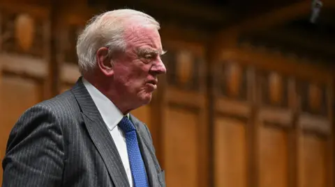 PA Media A man with white hair, combed back, stands to address the House of Commons with wood panels behind him. He is wearing a dark pin-striped suit jacket, white shirt and blue tie.