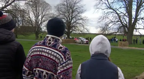 The photo shows a car driving through a park, on a road. It is avoiding hay bales, which are in the way as obstacles. There are a number of people watching on in the distance. There are three people in the front of the photo, wearing hats and hoods, watching on.