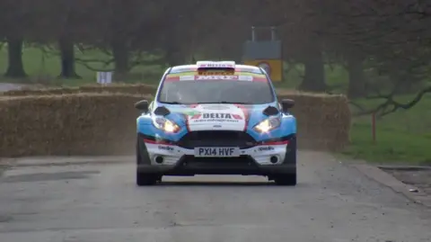 BBC The photo shows a rally car driving on a road, in a park. The car, which is a Ford, has been decorated with blue, white, red and black stickers. Behind it are hay bales, grass and trees. 