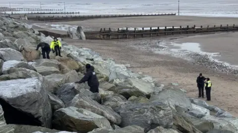 Phillip Norton/BBC Police officers clamber over rocks, placed as a sea defence, in front of a promenade. 