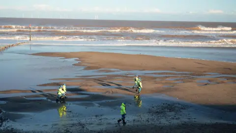 BBC/Joe Bilton People wearing high vis weather gear are walking across the beach. The tide is out.