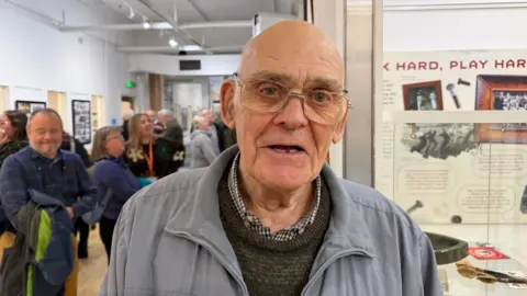Becki Bowden / BBC An elderly man wearing a shirt, brown jumper and a blue coat. He is bald and is wearing glasses. He is smiling at the camera. More people can be seen behind him looking at exhibits, which include a poster reading "Work hard, play hard".