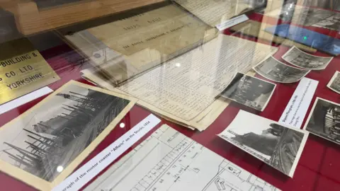 Becki Bowden / BBC A collection of black and white photographs, books and documents are laid out on red cloth in a glass cabinet.
