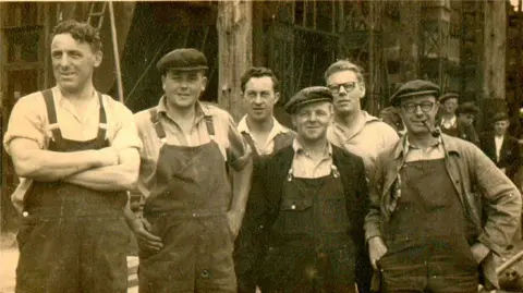 Brian Masterman Collection A black and white photograph of six workers at the Goole Shipyard. The men are wearing overalls and are smiling at the camera. Three of them are wearing flat caps and one is smoking a pipe.