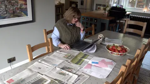 A woman with glasses and dark hair, wearing a brown knitted jumper is sitting indoors at a kitchen table with a laptop and newspapers in front of her. There is a bowl of red apples on the table and an A3-sized map with areas in pink - possibly a proposed solar farm location.