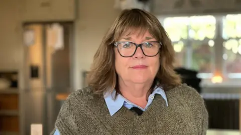 BBC A woman with glasses and dark hair, wearing a brown knitted jumper is sitting indoors in front of a window. 
