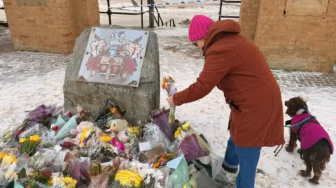 Jasmine Lowe/BBC A mount of flowers of various colours lay in the snow in front of two large castellated towers on a seafront. In the background is the sea. A woman wearing a red coat is placing flowers. She has a brown springer spaniel dog on a lead next to her.