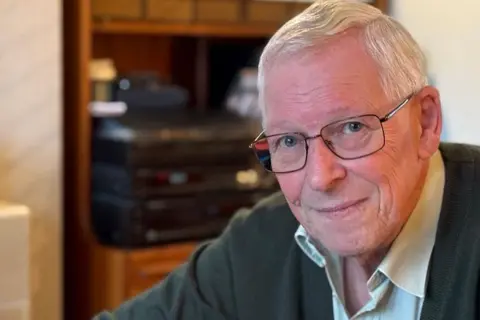 A man with short silver hair and dark-rimmed glasses sits in a living room in front of wooden shelves with a black hi-fi. He is wearing a green cardigan over a light shirt.