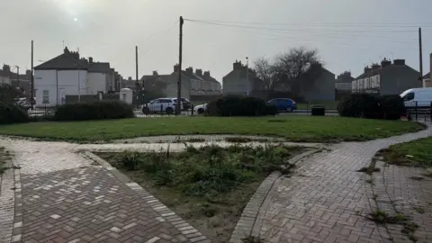 A small park, which looks a little neglected, with two paved paths curving to the left and right and patches of grass in between. Rows of terraced houses and another patch of grass can be seen in the background. Several cars are parked on a road in the middle-distance under a wintry grey sky.