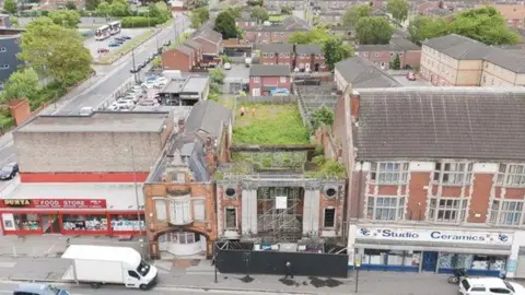 A drone view of the ruins of a bombed-out cinema, its early 20th century facade, which features colonnades and round windows, still standing. Behind the facade is a long grassy space. To either side are buildings dating from the same era, while a squat red food store can be seen to the far left. A white van and a car are parked on a road in the foreground. A modern housing estate can be seen in the background.