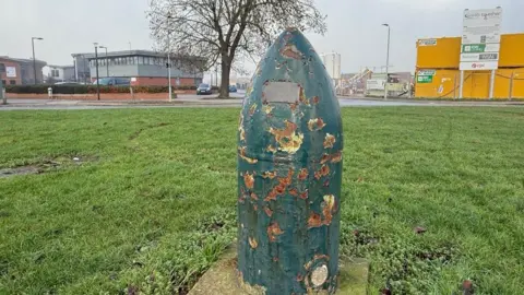 BBC A large green bullet-shaped bomb, covered in patches of rust, with a bronze plaque near the tip. It stands on a concrete base on a large patch of green grass in front of a road. In the background, a modern two-story building, a tall bare tree and a building site with yellow cabins can be seen.