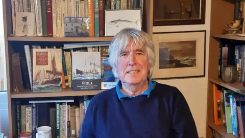 Dr Robb Robinson sits in his office surrounded by books on shelves. He has white hair and is wearing a navy blue jumper with a light blue shirt