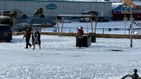 Kevin Shoesmith/BBC A couple, with their young child in a harness attached to the mother, walk through snow along a promenade. The dad is carrying a child's spade. 