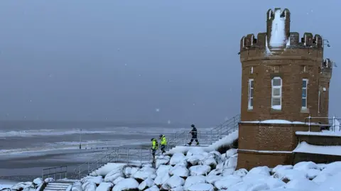 Kevin Shoesmith/BBC Three coastguard officials, two of them in yellow hi-vi jackets, make their way down a set of steps on to a snow-covered beach.