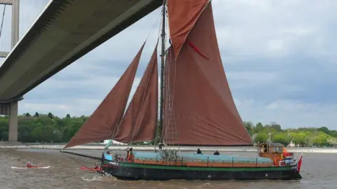 Mal Nicholson Spider T sailing under the Humber Bridge, It has red-coloured sails and rigging. There is a person in a canoe nearby.