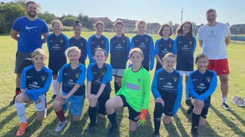 supplied A girl's football team pose in two rows. They are wearing blue strips and two male coaches stand smiling at each side.