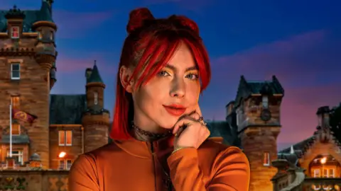 PA Media Traitors star Jessie Stride stood in front of the Traitors castle with flames in the background. She is wearing a bright orange top and has her red hair up in two buns.