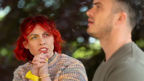PA Media The Traitors contestant Jessie Stride is seen chatting to fellow faithful Matthew. Jessie has bright red hair, yellow and orange eyeshadow and is wearing a rainbow jumper. The background has trees.