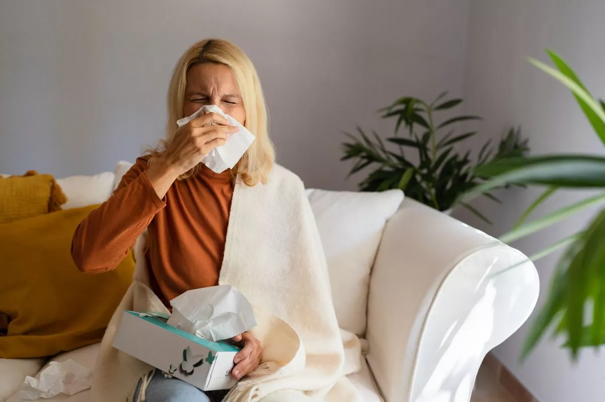 Sickwoman wrapped in blanket sitting on sofa and blowing her nose with a tissue