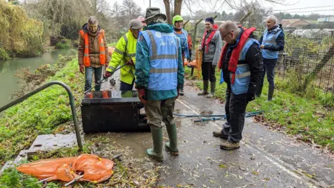Grantham RiverCare A group of litter pickers wearing blue, yellow and orange high vis vests, standing on tarmac path on top of a grassy embankment by the side of a river. They appear to have retrieved an old electrical appliance which is lying on the path with a length of blue rope attached to it.