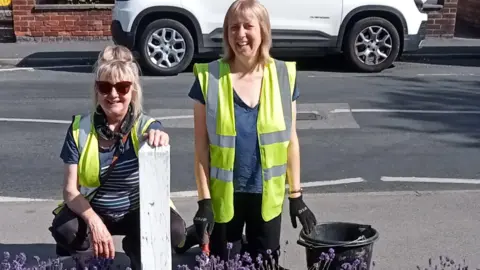 Beverley Urban Gardeners Two women smiling on a sunny day in a residential street with lavender growing in front of them. They are both wearing yellow hi vis vests. The woman on the left is squatting with her hand on a short white wooden post and the woman on the right is kneeling down, wearing black gloves with one hand resting on a black bucket.