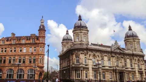 Getty Images Two old, large buildings stand next to each other. The one on the right is grand in design and has three silver domes on the top of turrets. The sky is blue with clouds. 