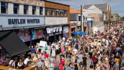 Humber Street Sesh Crowds of people enjoy drinks in sunny weather as part of the Humber Street Sesh music festival in Hull. They are gathered on a street with a row of shops and bars.