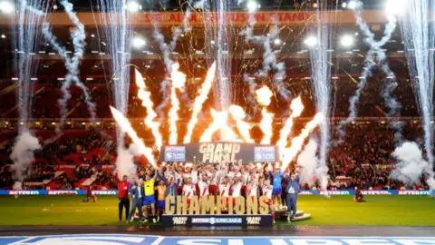 PA Media Hull KR celebrate winning the Super League Grand Final - the team all cheer in front of the stands while holding up the trophy as fireworks go off in the background.