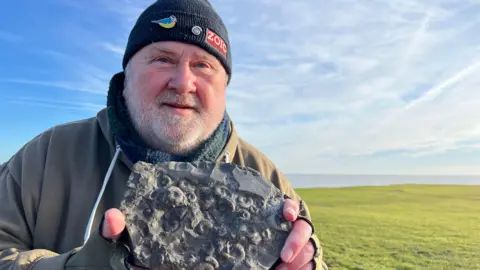 BBC A man wearing a black beanie hat with pin badges smiles into the camera. He is holding a dark, textured rock containing lots of circular fossils. He is standing on a grassed area under a bright, blue sky. The sea is visible in the background.