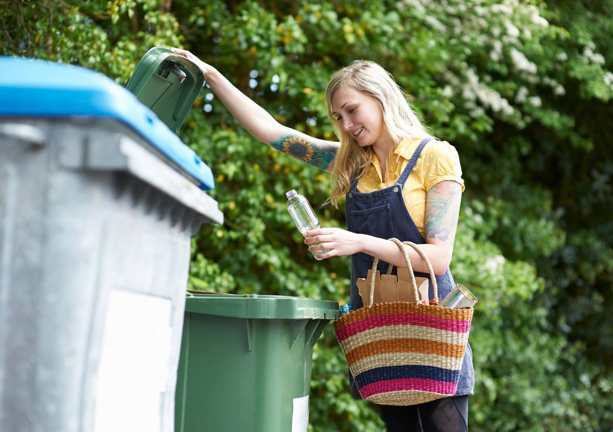 A happy young woman carrying a bag of recycling opens a bin to put a plastic bottle.