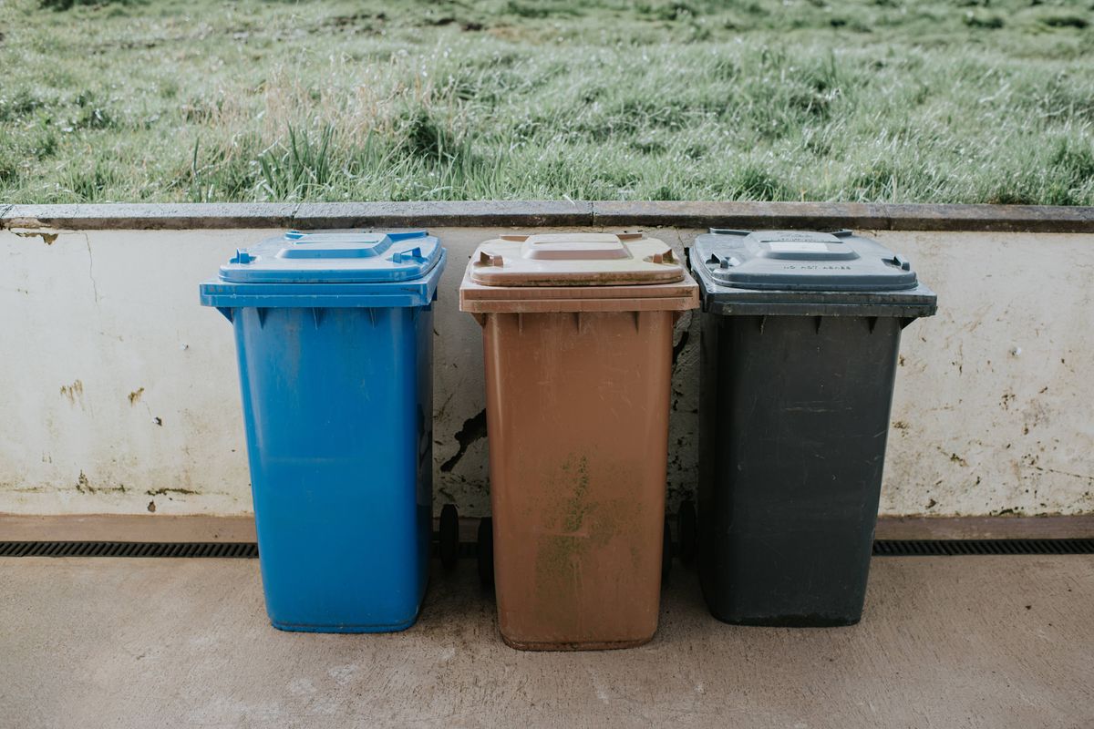 A brown, blue and black wheelie bin outdoors in front of a white wall.
