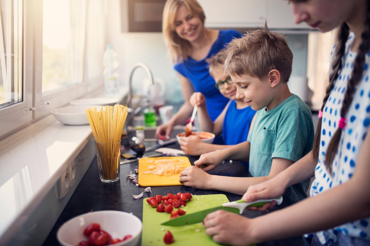 Three kids are cooking today! Mother is overseeing as a teenage girl and her two brother are chopping and mixing ingredients for family lunch.