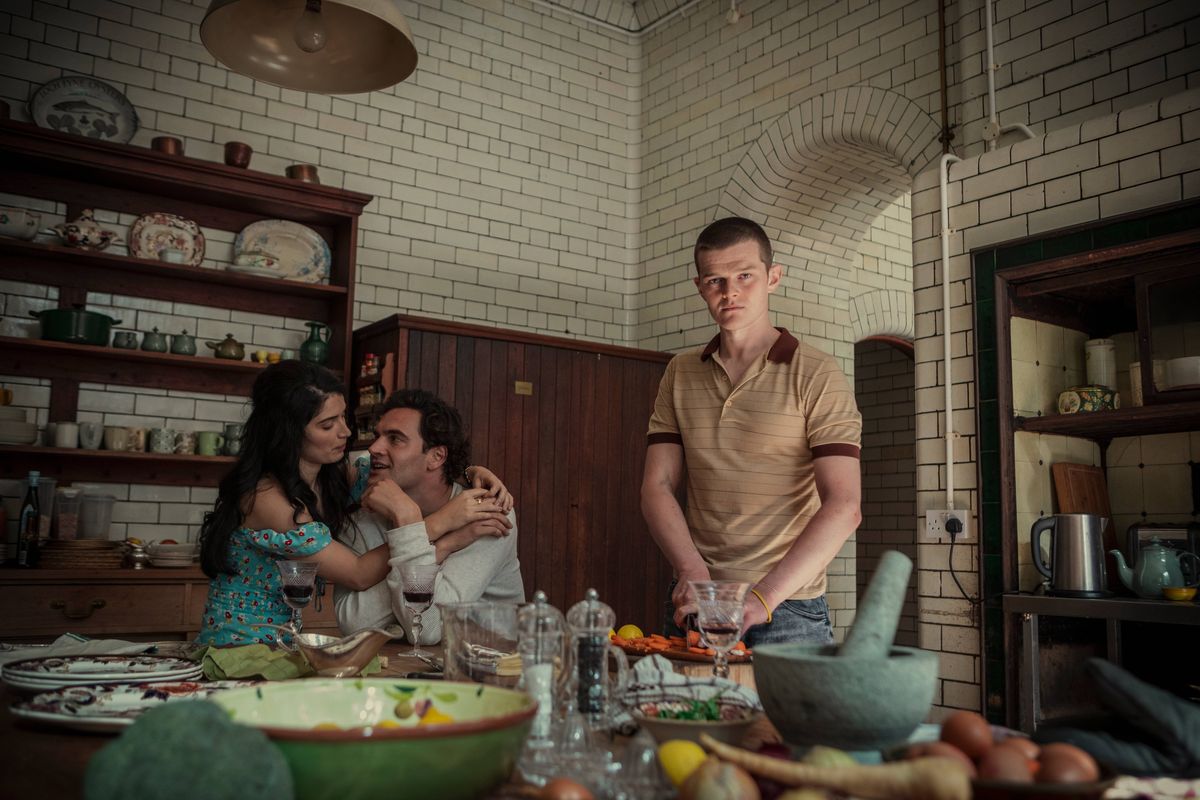 In a kitchen, a group of individuals are gathered around a table filled with various culinary items and ingredients. A man in a light-colored shirt stands at the table, actively engaged in food preparation, while others observe and interact nearby. The setting includes a wooden cabinet adorned with shelves, holding an assortment of dishes and kitchenware.