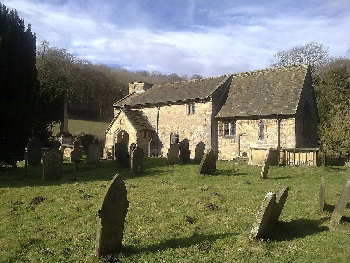 An old church with a gray roof is situated in the middle of a vast, green graveyard, surrounded by trees and a blue sky.