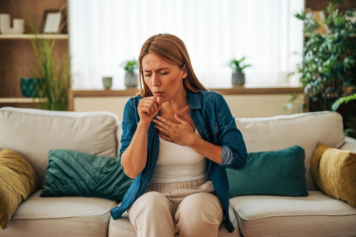 Mid adult woman suffering from cough, cold or flu, sitting on sofa at home, holding hand on chest, feeling unwell, experiencing symptoms of respiratory illness