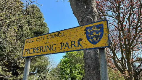 Richard Madden / BBC The yellow Hull City Council Pickering Park sign outside the main gate with the city's crest in blue there are trees behind it 