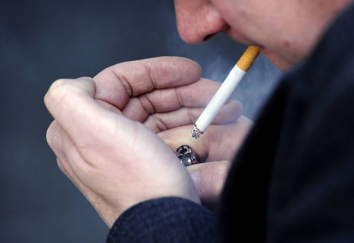 An individual is engaged in the act of smoking a cigarette, with the hand holding the cigarette raised to the mouth. The individual is dressed in a dark-colored long-sleeve shirt.